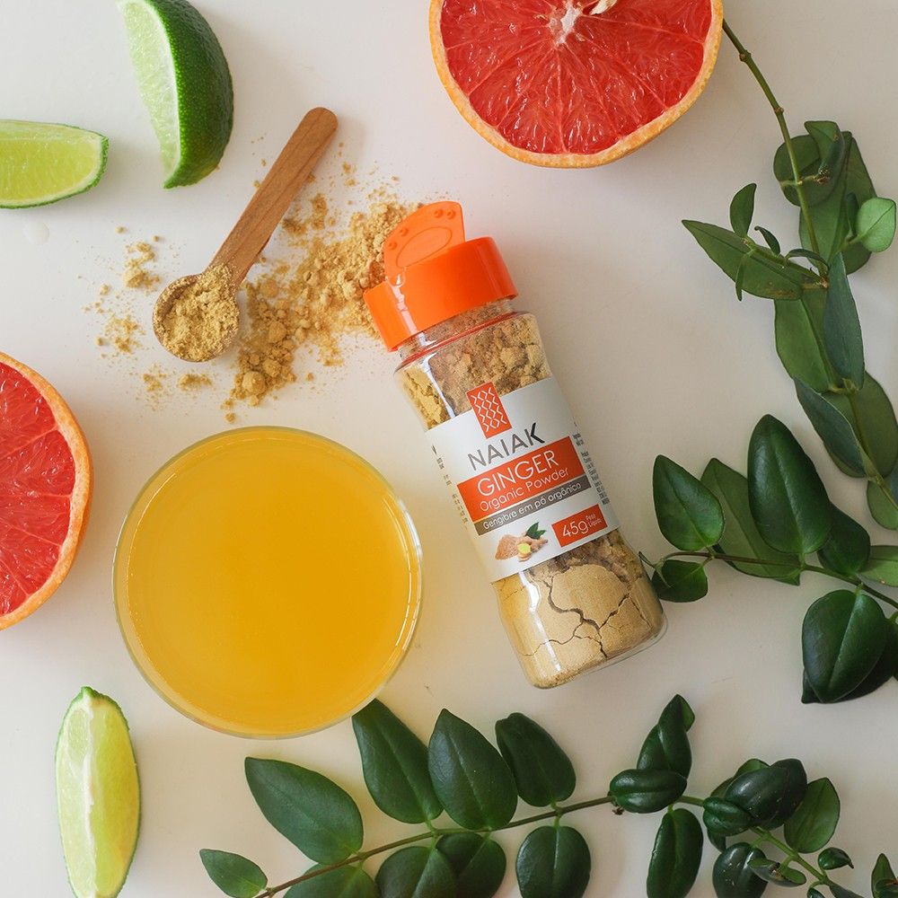 Pote de Gengibre em Pó Orgânico entre frutas cítricas, um copo de suco e uma colher com o pó